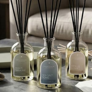 May include: Three glass diffuser bottles with black reeds. Each bottle has a label with text: "Amalfi Harmony", "Sea Salt Song", and "Vanilla Sonnet". The bottles are filled with a light yellow liquid.