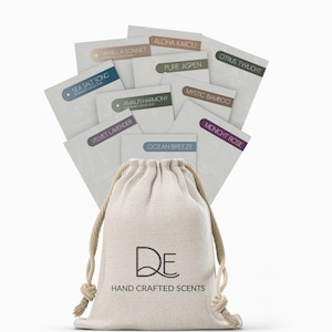 May include: A beige canvas bag with the text "DE HAND CRAFTED SCENTS" printed on the front. The bag is filled with small cards, each with a different scent name printed on it, such as "Aloha Kakou", "Pure Aspen", and "Sea Salt Song".