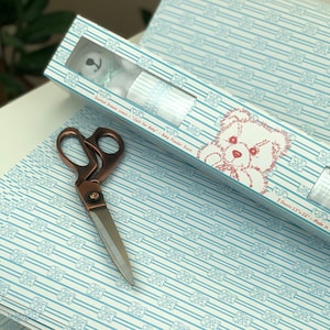 May include: A roll of white wrapping paper with a blue and white striped pattern and a repeating image of a teddy bear. The paper is packaged in a cardboard tube with a window showing a teddy bear and the text "Printed Design Liner - Satin for Baby - Baby Powder Scent".