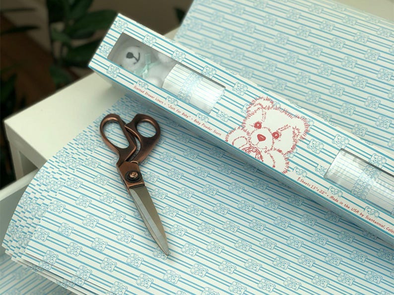 May include: A roll of white wrapping paper with a blue and white striped pattern and a repeating image of a teddy bear. The paper is packaged in a cardboard tube with a window showing a teddy bear and the text "Printed Design Liner - Satin for Baby - Baby Powder Scent".