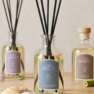 May include: Three glass diffuser bottles with black reeds. One bottle has a label that reads "Ocean Breeze" and another reads "Mystic Bamboo." The third bottle has a label that reads "Velvet Lavender." A seashell and bamboo stalks are also visible.
