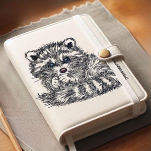 Pode incluir: Um diário branco com um desenho de guaxinim bordado. O guaxinim tem olhos azuis e está a segurar a pata na boca. O diário tem uma faixa elástica branca e um fecho de botão dourado. Uma fita de marcador branca é visível.