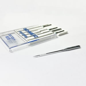 May include: A clear plastic case containing a pack of six silver sewing needles. The needles are labeled "SCHMETZ UNIVERSAL" and "130/705 H 60/8". A single silver sewing needle is lying on a white surface.