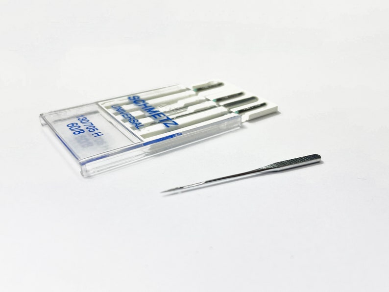 May include: A clear plastic case containing a pack of six silver sewing needles. The needles are labeled "SCHMETZ UNIVERSAL" and "130/705 H 60/8". A single silver sewing needle is lying on a white surface.