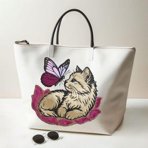 May include: Cream tote bag with black handles and a zip closure. It features an embroidered design of a tan cat with blue eyes and a pink and purple butterfly. Sunglasses are at the bottom.