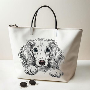 Puede incluir: Un bolso tote de lona blanca con una ilustración en blanco y negro de un perro salchicha de pelo largo con ojos azules. El bolso tiene asas negras y cierre de cremallera. Unas gafas de sol redondas están delante del bolso.