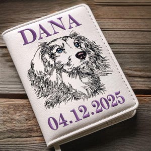Könnte beinhalten: Weißes Notizbuch mit einer detaillierten Stickerei eines Hundegesichts mit blauen Augen, dem lila Schriftzug "DANA" und dem Datum "04.12.2025". Das Notizbuch hat einen genähten Rand.