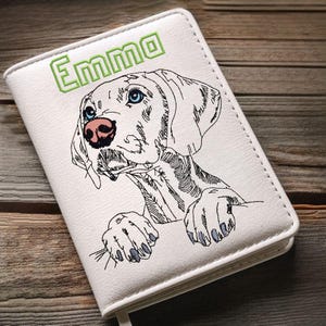 May include: White notebook with an embroidered dog illustration. The dog has blue eyes and a pink nose. The name "Emma" is embroidered in green above the dog. The notebook is closed and has a white border.