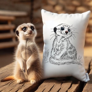 May include: A white pillow with a black and white line drawing of a meerkat sitting up. The meerkat has a cute expression and is looking to the right. A real meerkat is sitting on a wooden surface in the background.