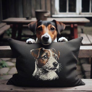 Op de afbeelding: Een rechthoekig donkergrijs sierkussen met een gedetailleerd geborduurd portret van een Jack Russell Terriër. Het gezicht van de hond is weergegeven in tinten wit, bruin en zwart, passend bij het uiterlijk van de echte hond die zijn kin op het kussen laat rusten.