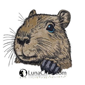 Könnte beinhalten: Ein Stickmotiv, das den Kopf eines braunen Capybaras mit schwarzen Stickdetails, einem leuchtend blauen Auge und einer sichtbaren dunklen Pfote zeigt. Darunter steht der Text LunaChic.com und Embroidery files Made in Germany.