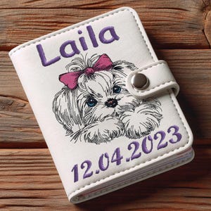 Pode incluir: Carteira branca de couro sintético com um laço rosa e uma ilustração de um cão branco. A carteira tem o nome "Laila" e a data "12.04.2023" bordada em roxo.
