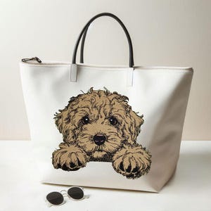 May include: A white canvas tote bag featuring a detailed illustration of a fluffy tan puppy with dark eyes and paws resting on the bottom edge. The bag has two black shoulder straps and a top zipper closure. A pair of black sunglasses sits below.