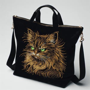 May include: Black tote bag featuring an embroidered cat face with green eyes and gold-colored fur. The bag has a zipper closure, black handles, and a detachable shoulder strap with gold-colored hardware.