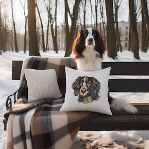 May include: A brown, white, and black dog sits on a park bench with a white pillow featuring a dog portrait. A second white pillow and a brown and cream plaid blanket are also on the bench. The background shows a snowy park.