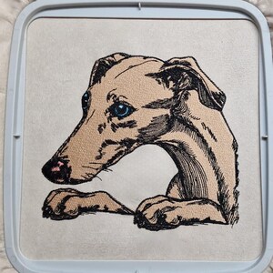 Könnte beinhalten: Ein Stickdesign eines beigefarbenen Windhundes mit schwarzen Linien und leuchtend blauen Augen, eingespannt in einen hellgrauen Kunststoff-Stickrahmen. Der Hund ist im Profil dargestellt, mit gedrehtem Kopf und nach vorne ruhenden Pfoten.