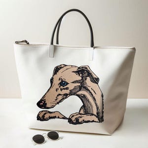 Könnte beinhalten: Eine weiße Canvas-Tragetasche mit einer detaillierten Illustration eines beigefarbenen Windhundes mit blauen Augen. Die Tasche hat zwei schwarze Griffe und einen Reißverschluss oben. Eine runde schwarze Sonnenbrille liegt vor der Tragetasche.