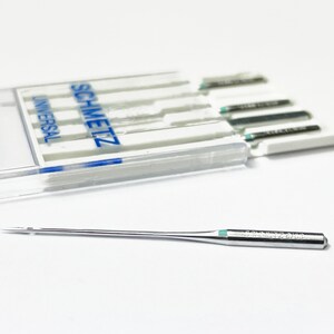 May include: A close-up of a single silver sewing needle with a green tip. The needle is labeled "SCHMETZ 12/80". The needle is lying on a white surface. A clear plastic case with a blue label that reads "SCHMETZ UNIVERSAL" is in the background.