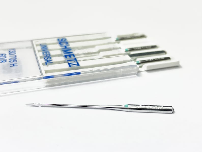 May include: A close-up of a single silver sewing needle with a green tip. The needle is labeled "SCHMETZ 12/80". The needle is lying on a white surface. A clear plastic case with a blue label that reads "SCHMETZ UNIVERSAL" is in the background.