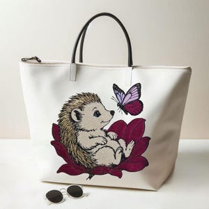 May include: White tote bag with a zipper closure and black handles. The bag features an embroidered design of a hedgehog sitting on a burgundy flower, with a purple butterfly nearby. Sunglasses are at the bottom.
