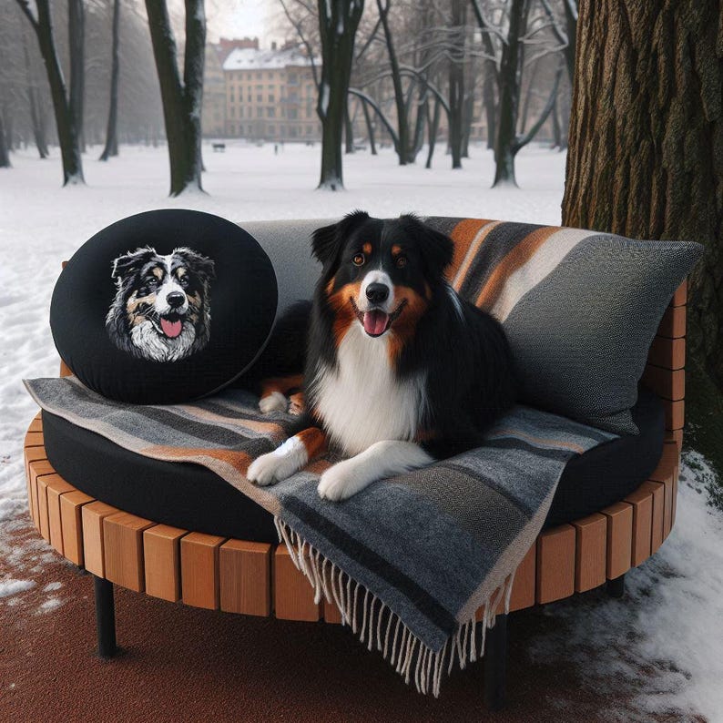 Stickdatei Australian Shepherd Sanzy als winterliches Anwendungsbeispiel, detailreich aufgestickt auf ein rundes, schwarzes Zierkissen, das neben einem echten, wunderschönen Australian Shepherd auf einer gemusterten Wolldecke auf einer runden Holzbank im verschneiten Park liegt von LunaChild.com