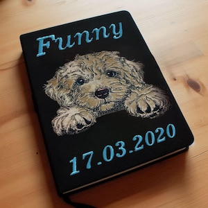 Può includere: Un taccuino nero con un cane bianco ricamato sul davanti. Il cane guarda la fotocamera con le zampe appoggiate sul bordo del taccuino. Il taccuino ha il nome "Funny" e la data "17.03.2020" ricamata sul davanti con filo blu.