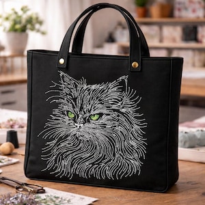 Könnte beinhalten: Schwarze Tragetasche mit einem gestickten Katzenkopf. Die Katze hat grüne Augen und weiße Details. Die Tasche hat zwei schwarze Griffe und goldfarbene Nieten. Das Fell der Katze wird durch aufwendige weiße Nähte dargestellt. Die Tasche ist aus robustem Material gefertigt.