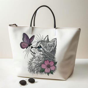 Könnte beinhalten: Weißer Shopper mit schwarzen Griffen und Reißverschluss. Die Tasche hat ein gesticktes Design einer Katze mit blauen Augen, einem lila Schmetterling und einer rosa Blume. Sonnenbrille im Vordergrund.