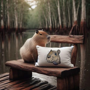 Könnte beinhalten: Ein rechteckiges, cremefarbenes Dekokissen mit einer detaillierten Illustration eines Capybara-Kopfes auf der Vorderseite. Das Kissen liegt auf einer rustikalen Holzbank neben einem lebenden Capybara vor einem unscharfen Wald- und Wasserhintergrund.