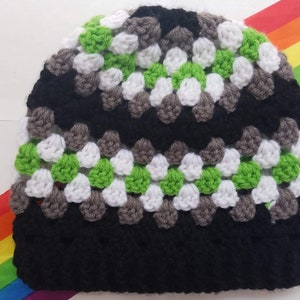 Op de afbeelding: Een zwarte, groene, witte en grijze gebreide beanie. De beanie heeft een textuurpatroon.