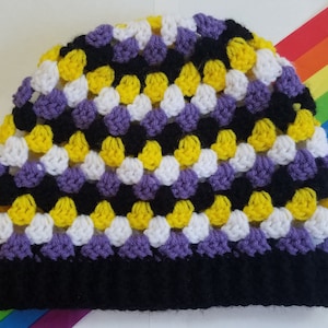 Può includere: Un berretto lavorato a uncinetto con un motivo di colore nero, giallo, bianco e viola. Il berretto è su uno sfondo arcobaleno.