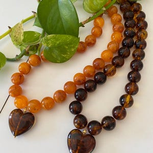 Könnte beinhalten: Zwei Halsketten mit Herzanhängern. Eine Halskette hat bernsteinfarbene Perlen und einen dunkelbraunen Herzanhänger. Die andere Halskette hat dunkelbraune Perlen und einen Herzanhänger. Grüne Blätter sind im Hintergrund.