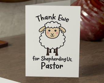 May include: A white greeting card featuring a cartoon sheep illustration with the black text "Thank Ewe for Shepherding Us, Pastor" printed above and below the sheep. A second folded white card is positioned in the foreground.