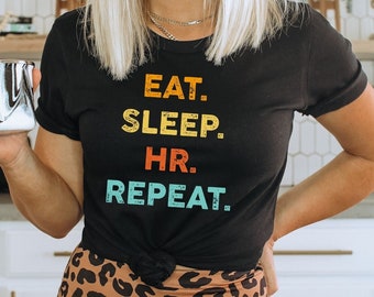 May include: A black t-shirt with the text "EAT. SLEEP. HR. REPEAT." in colorful retro-style lettering. The person is wearing leopard print leggings.