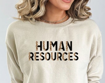 May include: A beige sweatshirt with the text "HUMAN RESOURCES" printed in black letters with a leopard print pattern.