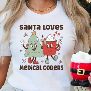 Puede incluir: Camiseta blanca con un diseño retro que presenta un árbol de Navidad y una taza de cacao, ambos con caras sonrientes. El texto "SANTA LOVES MEDICAL CODERS" está impreso encima y debajo del gráfico. Se sostiene una taza con temática de Papá Noel.