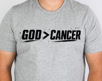 May include: A heather gray short-sleeved t-shirt featuring the black text GOD > CANCER printed across the chest. The graphic includes distressed, brush-stroke style accents surrounding the lettering on a plain crew neck garment.