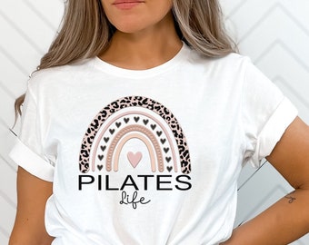 May include: White t-shirt with a pink and brown rainbow design with black leopard print accents and black hearts. The text "PILATES life" is printed below the rainbow.