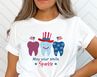 May include: White t-shirt with a graphic of three teeth wearing red, white, and blue patriotic hats and the text "May your smile Sparkle".