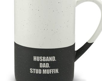 May include: A tall ceramic mug with a two-tone design, featuring a white speckled upper half and a solid black speckled lower half. The text HUSBAND. DAD. STUD MUFFIN. is printed in white on the black section. It has a curved handle.