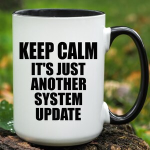 May include: A white ceramic mug with a black interior and a black handle. The front of the mug features the black printed text: KEEP CALM IT'S JUST ANOTHER SYSTEM UPDATE. The mug has a cylindrical body and a curved handle on the right side.