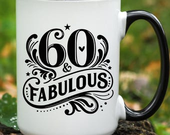 May include: A white ceramic mug with a black interior and handle. The front features black decorative typography that reads 60 & FABULOUS, accented with swirls, small stars, and a tiny heart centered between the numbers.