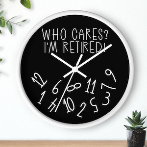 May include: A black and white wall clock with a white rim. The clock face has a black background with white numbers and hands. The text "WHO CARES? I'M RETIRED!" is written in white on the clock face.