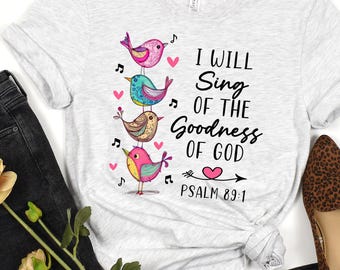 May include: A light gray heather t-shirt featuring a stack of four colorful illustrated birds, musical notes, and small pink hearts. The text reads: I WILL SING OF THE GOODNESS OF GOD PSALM 89:1. The shirt is paired with black jeans and leopard print flats.