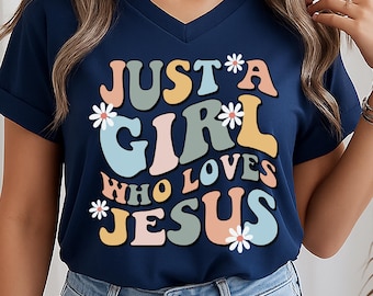 May include: A navy blue V-neck t-shirt with short sleeves and rolled cuffs. The front features the text "JUST A GIRL WHO LOVES JESUS" in a multi-colored, retro-style font, accented with small white daisy illustrations scattered around the lettering.