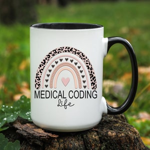 May include: White ceramic mug with a black handle. The mug features a pink and black rainbow with hearts and the text "MEDICAL CODING life".