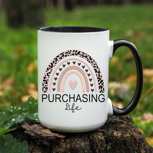 May include: White ceramic mug with a black interior and handle. The mug features a rainbow design with leopard print, hearts, and the words "PURCHASING Life" in black script. The mug is sitting on a tree stump.
