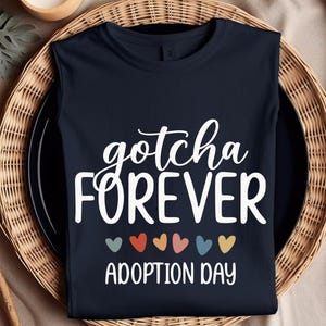 Peut inclure: T-shirt bleu marine avec l'inscription blanche "gotcha FOREVER ADOPTION DAY". En dessous, six cœurs colorés. Le t-shirt est présenté sur une assiette noire à l'intérieur d'un panier tressé.