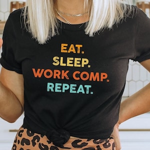 May include: A black t-shirt with the text "Eat. Sleep. Work Comp. Repeat." in a retro font with colorful accents. The shirt is worn by a person with blonde hair and a leopard print skirt.