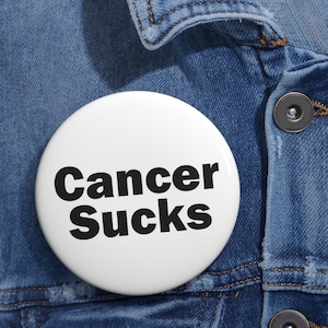 May include: A white, round button with the words "Cancer Sucks" in bold, black letters. The button is pinned to a blue denim jacket, with visible stitching and a button closure.